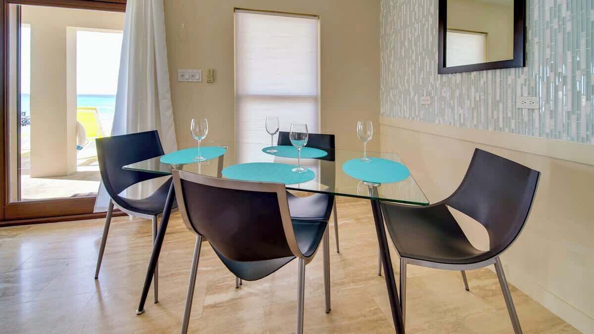 Un coin repas avec une table en verre et quatre chaises, une fenêtre avec des stores et un miroir au mur.