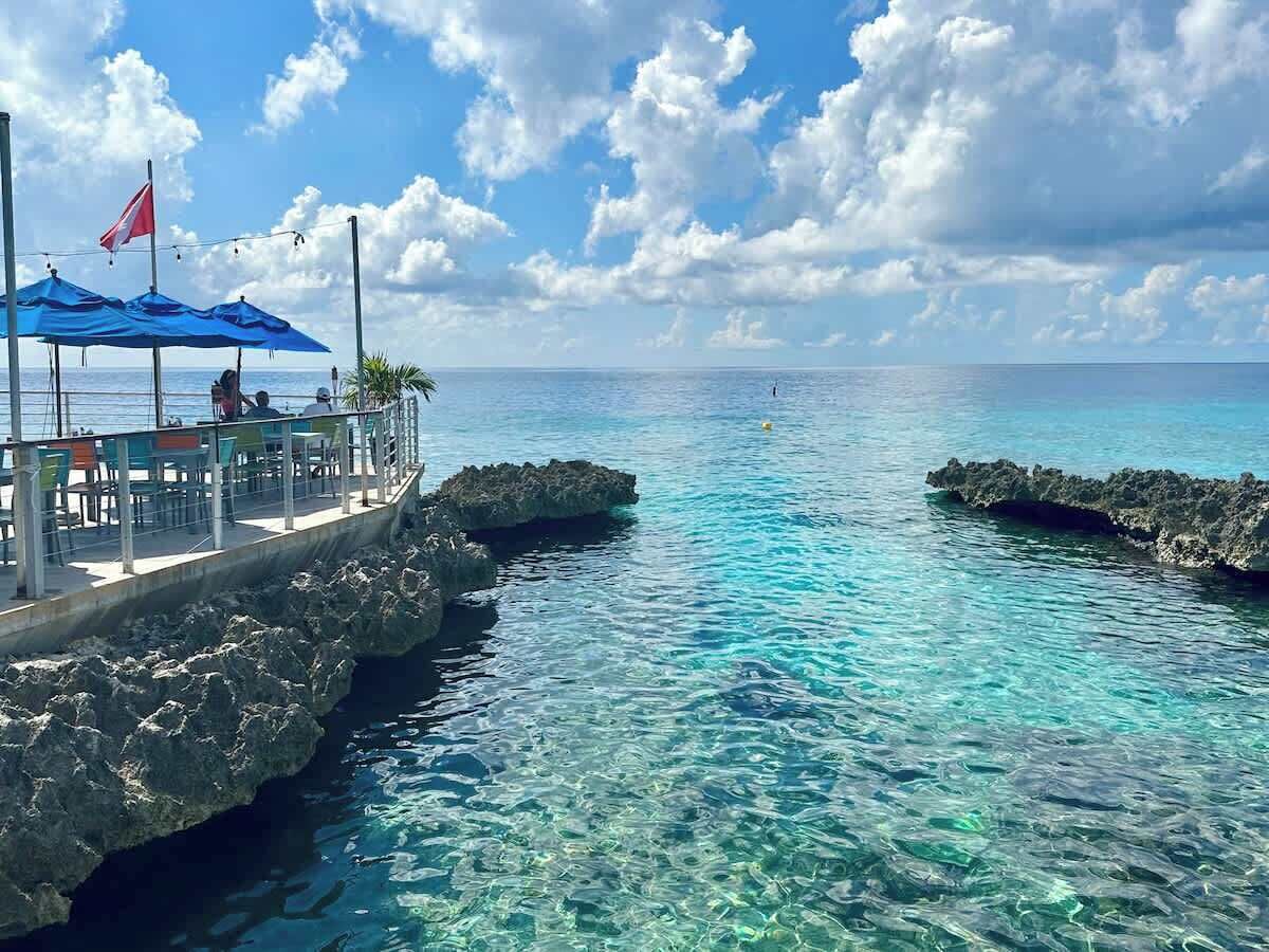 Un muelle con mesas y sillas, con vista a aguas turquesas cristalinas y formaciones rocosas.