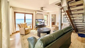 A modern living room with a large TV, a green sofa, and a balcony with a view of the sea. - Seven Mile View #1 - 2 BR Oceanfront Condo & Pool (West Bay)