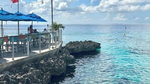 A seaside restaurant with outdoor seating, a flag, and a rocky outcrop in the water. - Seven Mile View #1 - 2 BR Oceanfront Condo & Pool (West Bay)