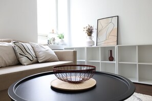 A modern living room with a round black table, a wire basket on a wooden coaster, a beige sofa with patterned pillows, and a white shelving unit with decorative items.