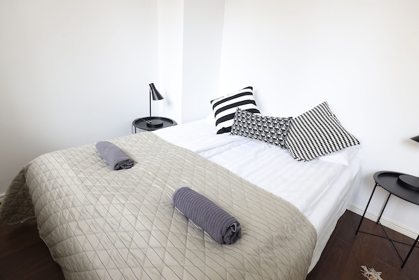 A neatly made bed with a quilted bedspread and two rolled towels, a black floor lamp, and two patterned pillows on a white mattress. - 2ndhomes Bright 1BR Home in Kamppi (Helsinki)
