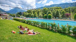 Ein Poolbereich mit Liegestühlen auf der Wiese, umgeben von Bäumen und einem Berg im Hintergrund.