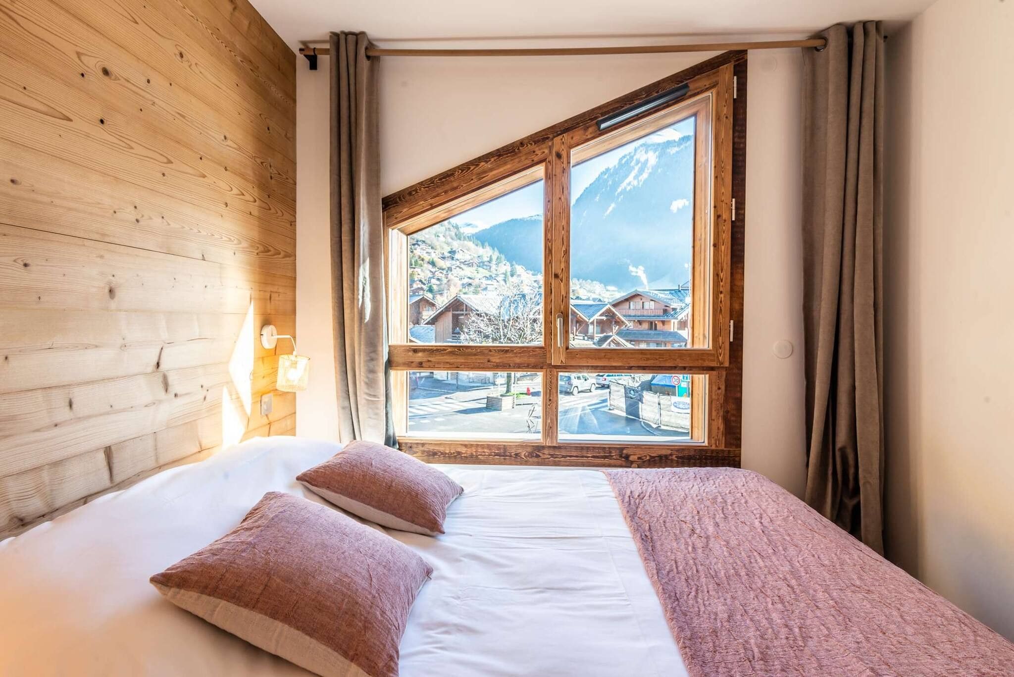 A bedroom with a large window, wooden walls, a bed with white bedding and two pillows, and a nightstand with a lamp.