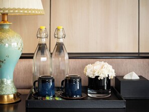 A tray with two glass bottles, a blue mug, a black mug, a vase with white flowers, a tissue box, and a green packet. - M'Gloria Cruise (Ha Long)