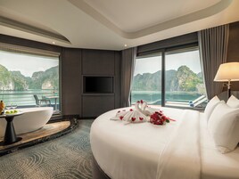 Habitación de hotel con una cama grande, televisión montada en la pared, zona de comedor con mesa y sillas, y vistas a un cuerpo de agua y montañas.