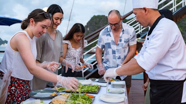 Un groupe de personnes prépare des aliments sur le pont d’un bateau, avec une rambarde et de la verdure en arrière-plan.