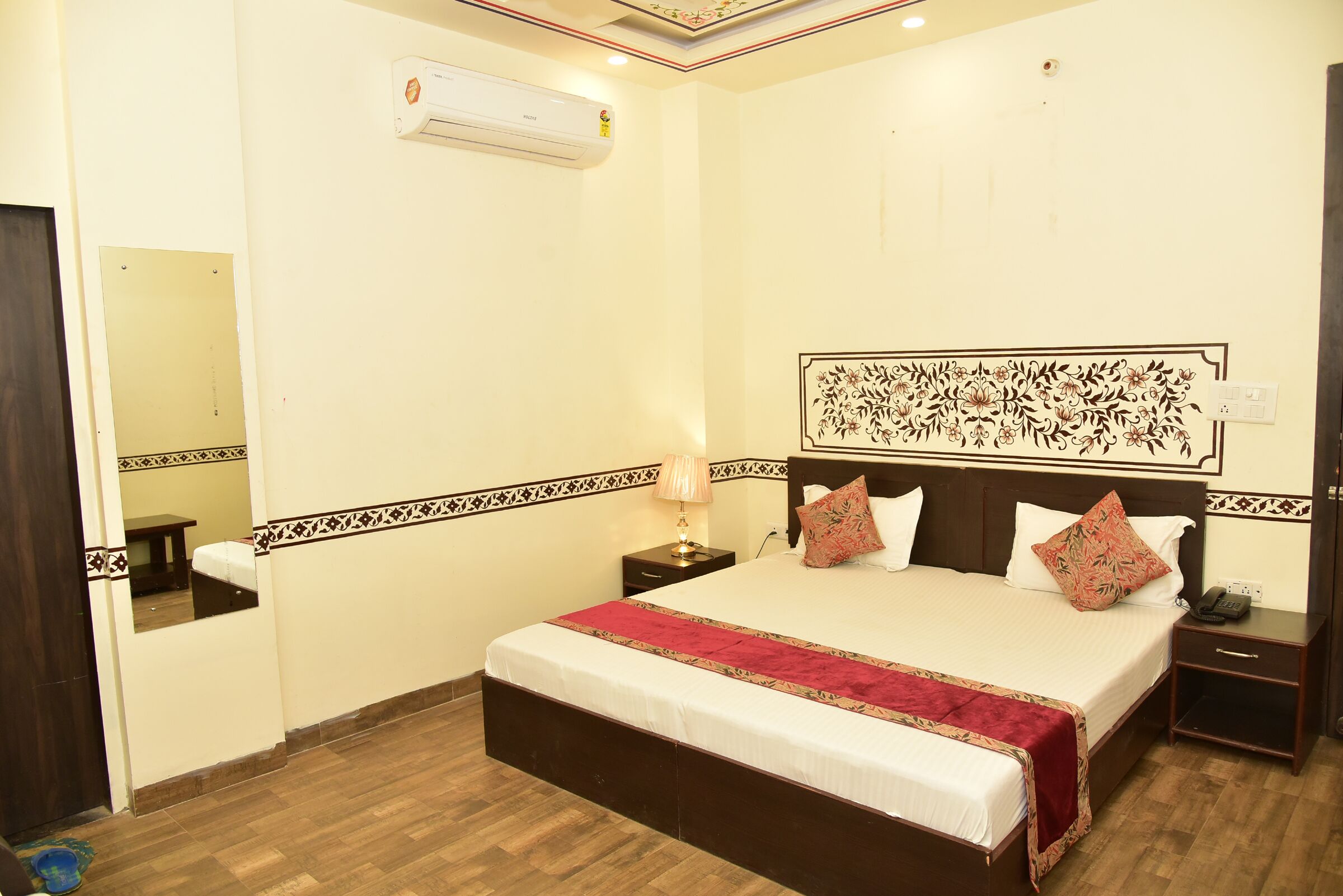 A neatly arranged hotel room with a large bed, a bedside table, a nightstand, a mirror, and an air conditioning unit on the wall.