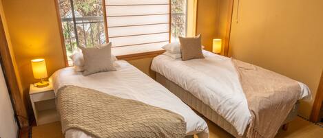 Una habitación con dos camas, cada una con almohada y manta, una mesita de noche con lámpara, y una ventana con vista a árboles.
