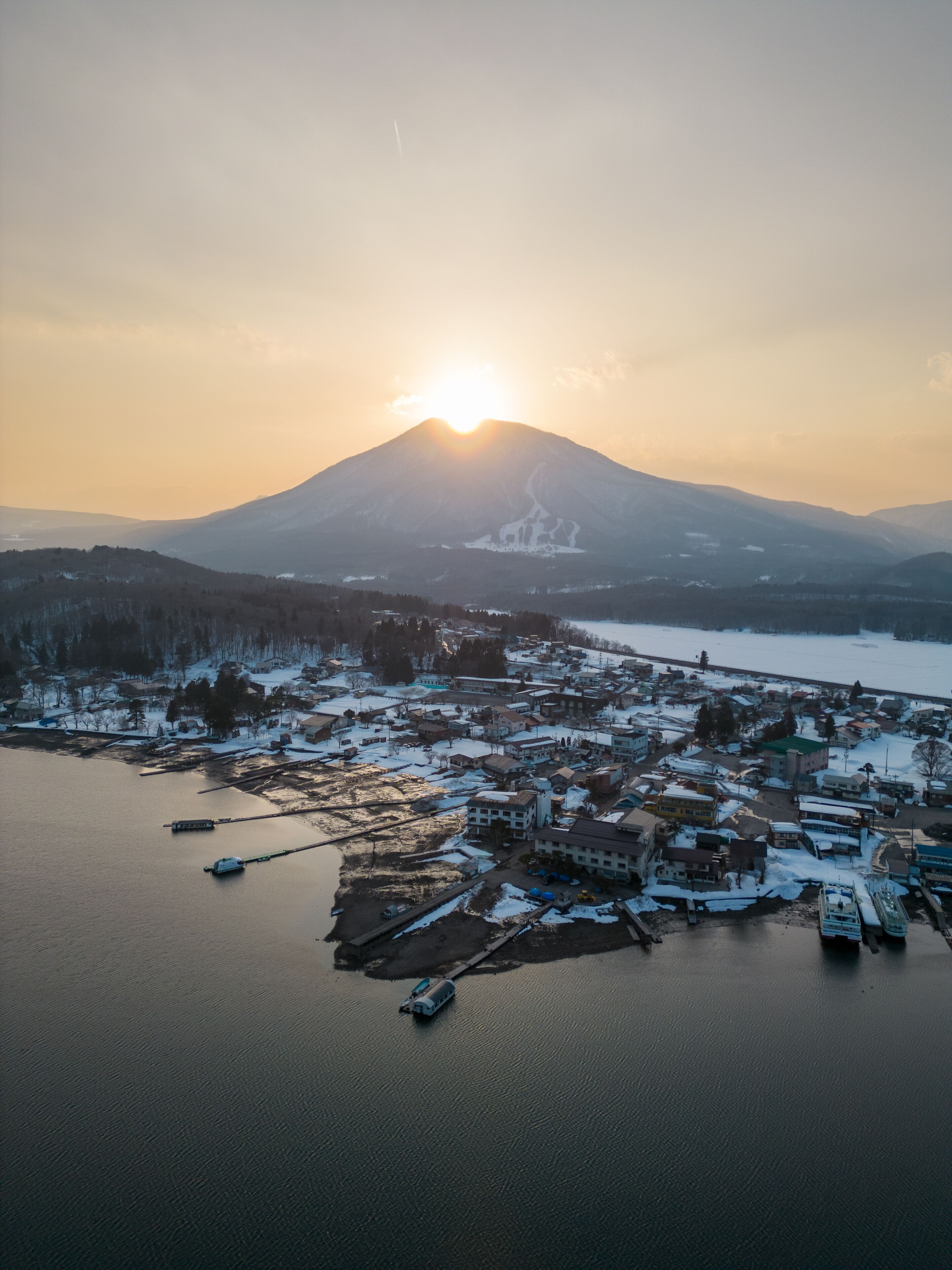 An aerial view of a coastal town with a mountain in the background, boats docked at the harbor, and snow-covered buildings.
