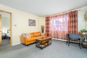 En stue med sofa, sofabord i træ, stol og udsigt gennem lette gardiner.