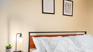 Una cama bien tendida con ropa de cama blanca, una mesita de noche con lámpara y planta en maceta, y dos obras de arte enmarcadas en la pared.