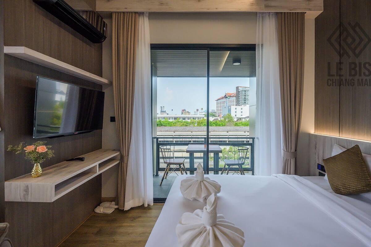 A hotel room with a bed, a TV, a balcony with a view of buildings, and a sign on the wall.
