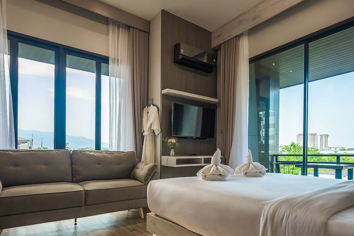 A modern hotel room with a large bed, a sofa, a TV, and a balcony with a city view.