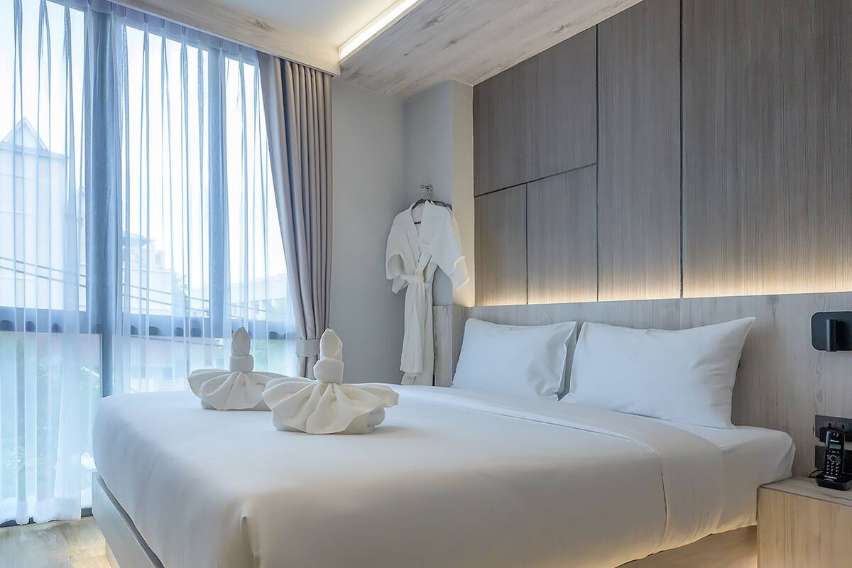A modern hotel room with a large bed, a bedside table with a phone, and a view of the outdoors through sheer curtains.