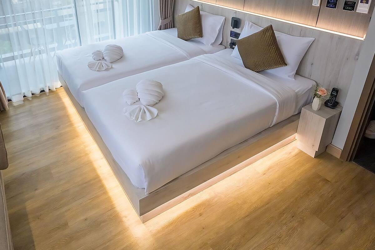 A hotel room with two beds, a wooden floor, a mirror, a robe, and a small table with a vase.