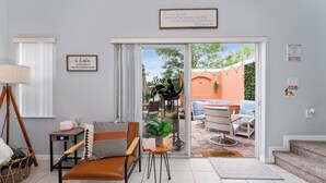 En hyggelig stue med en sofa, et lille bord og en stol. Der er en skydedør, der fører ud til en udendørs terrasse med bord og stole.