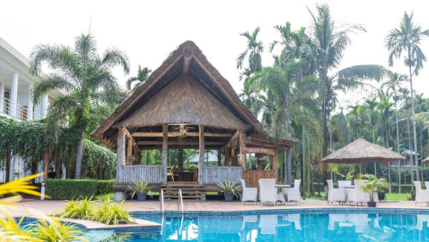 A pool area with a thatched-roof gazebo, white lounge chairs, and palm trees. - Zone Niathu Dimapur By The Park (Dimapur)