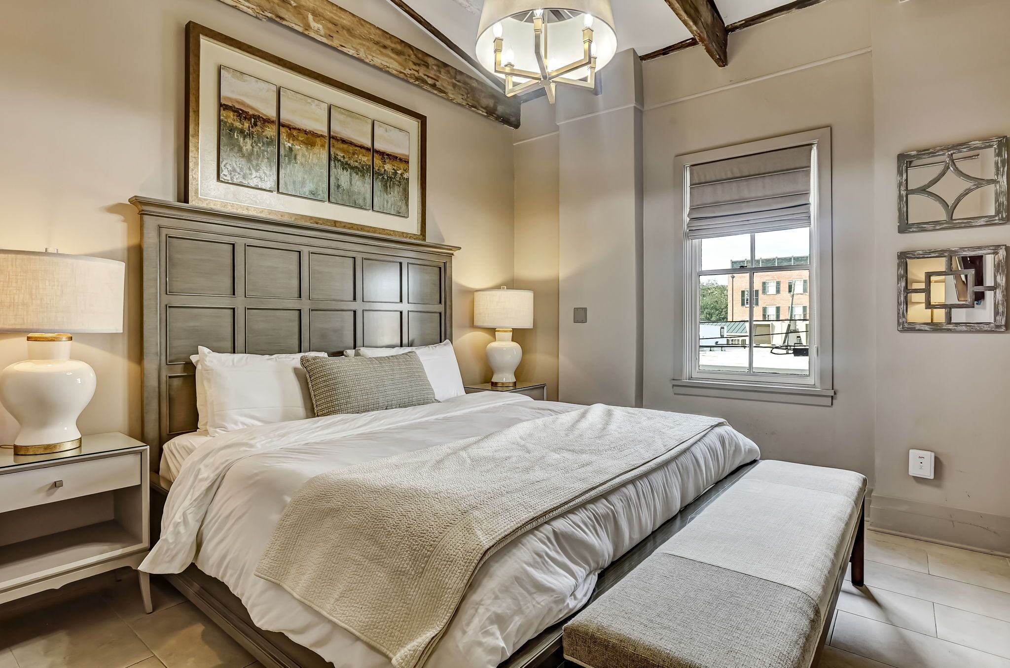 A bedroom with a large bed, two bedside tables with lamps, a headboard, a window with a view, and a chandelier.