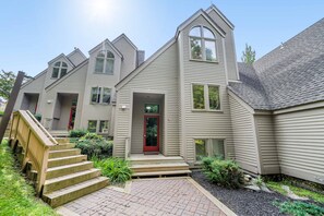 Une maison à deux étages avec une porte rouge, un porche à l’avant et un escalier en bois menant à l’entrée.