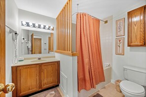 Une salle de bain avec un meuble-lavabo en bois, un miroir avec éclairage intégré, un rideau de douche, des toilettes et des tableaux encadrés accrochés au mur.