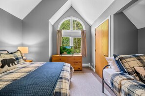 Une chambre avec un plafond en pente, une grande fenêtre avec des rideaux, un lit recouvert d’une couette bleue, une table de chevet avec une lampe et une commode.
