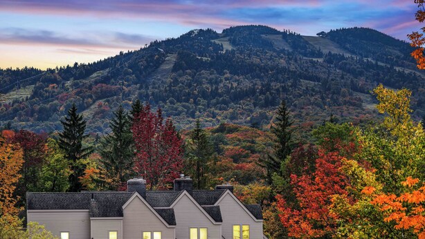 Une maison à deux étages, dotée de nombreuses fenêtres, entourée d’un feuillage automnal et avec une chaîne de montagnes en arrière-plan.