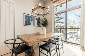 A dining area with a wooden table, black chairs, a chandelier, framed artwork, and a view of a snowy landscape through large windows.