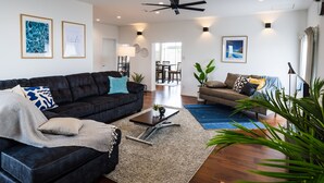 A modern living room with a dark sofa, a coffee table, and a blue rug. There are two framed pictures on the wall, a ceiling fan, and a dining area in the background. - MARINUS (Miyakojima)
