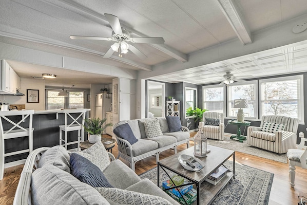 A spacious living room with a grey sofa, striped armchairs, and a coffee table. There are ceiling fans, a kitchen area in the background, and large windows providing natural light.