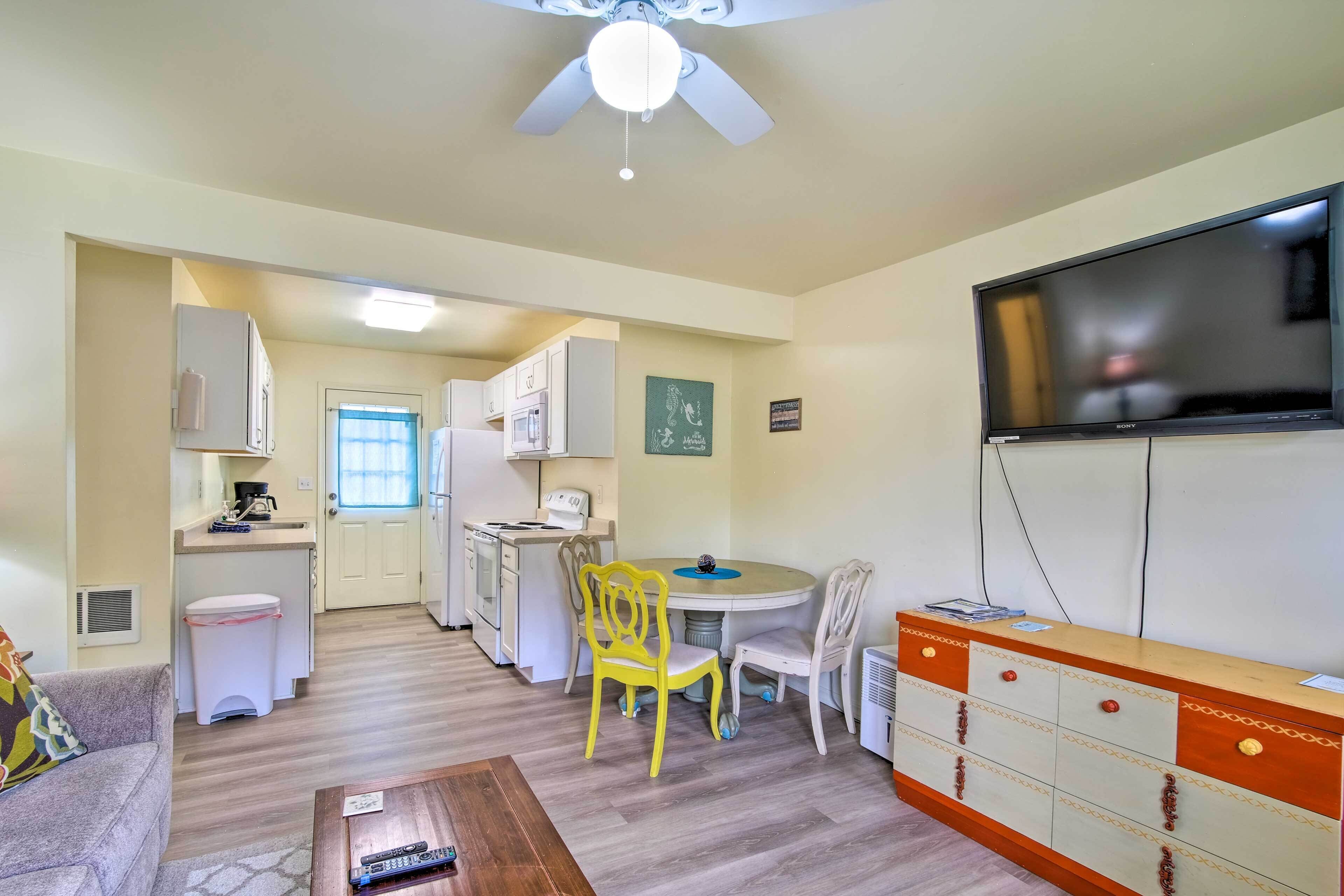 A compact living space with a kitchenette, a dining area with yellow chairs, a TV mounted on the wall, and a ceiling fan.