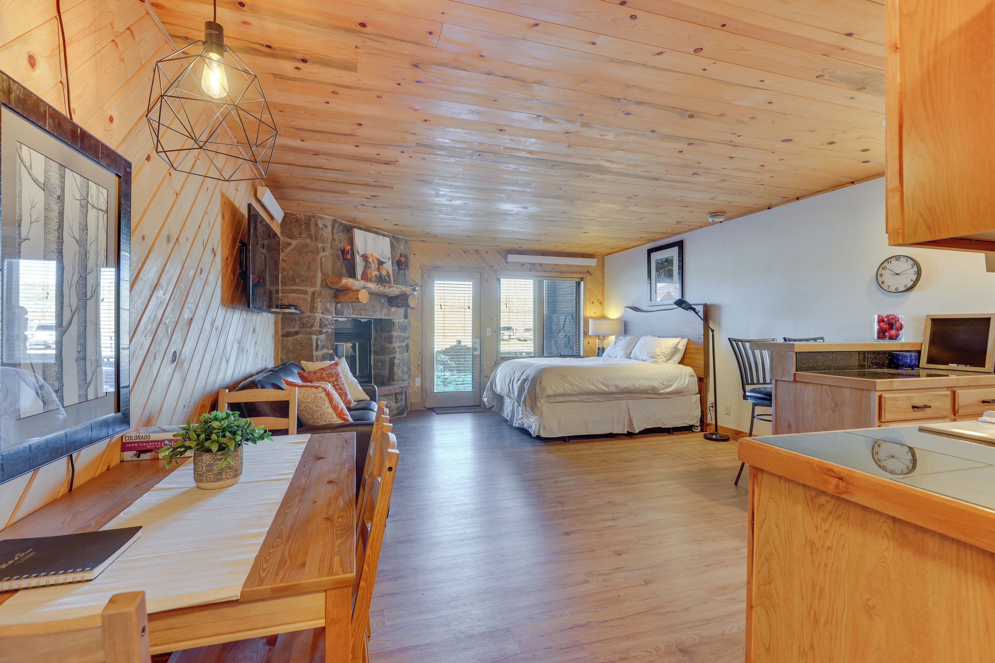 A cozy cabin interior with a wooden ceiling, a dining area with a bench, a bed, and a kitchen area with a clock.