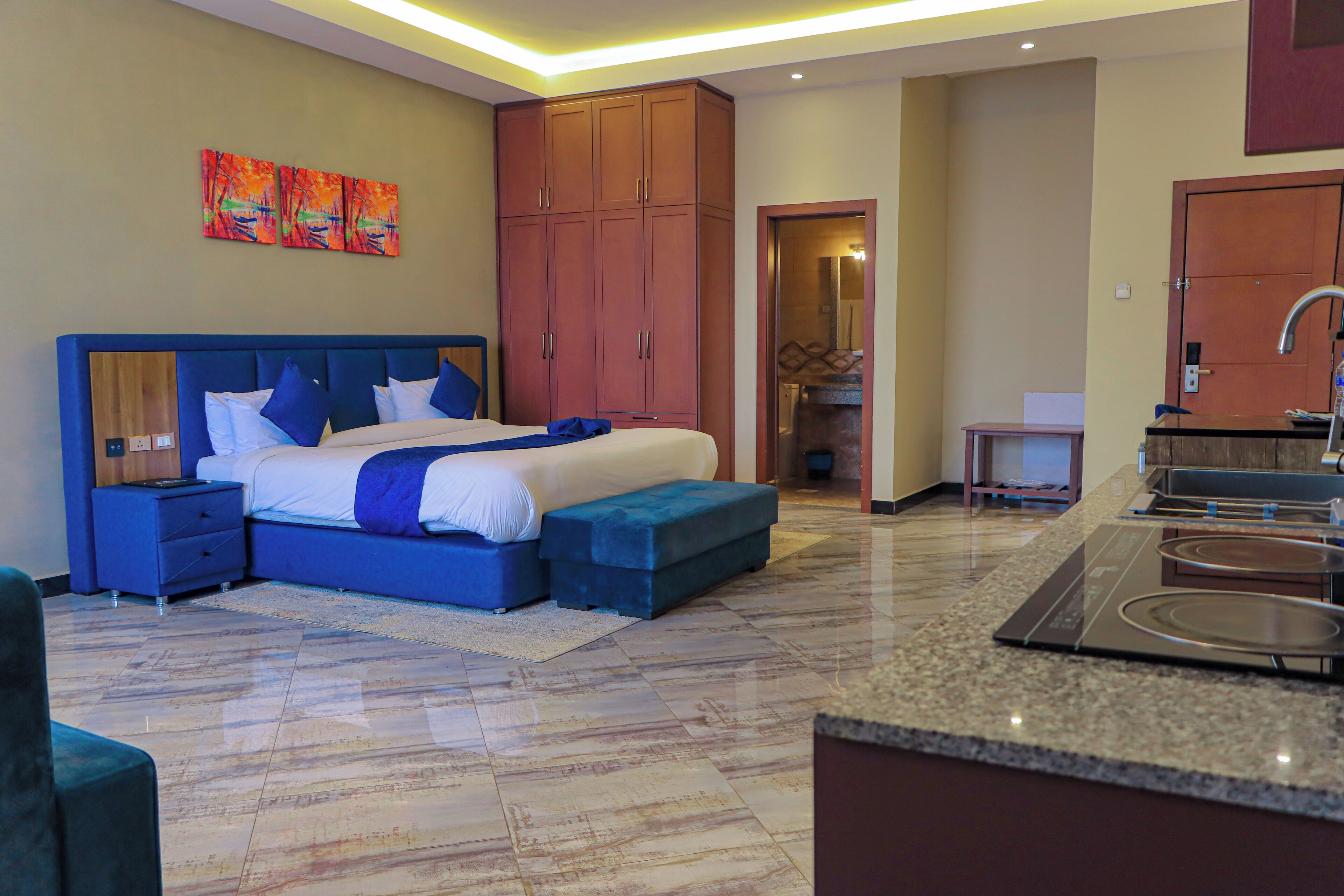 A modern hotel room with a large bed, a built-in desk, a kitchenette, and a bathroom visible through an open door.