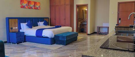 A modern hotel room with a large bed, a built-in desk, a kitchenette, and a bathroom visible through an open door.