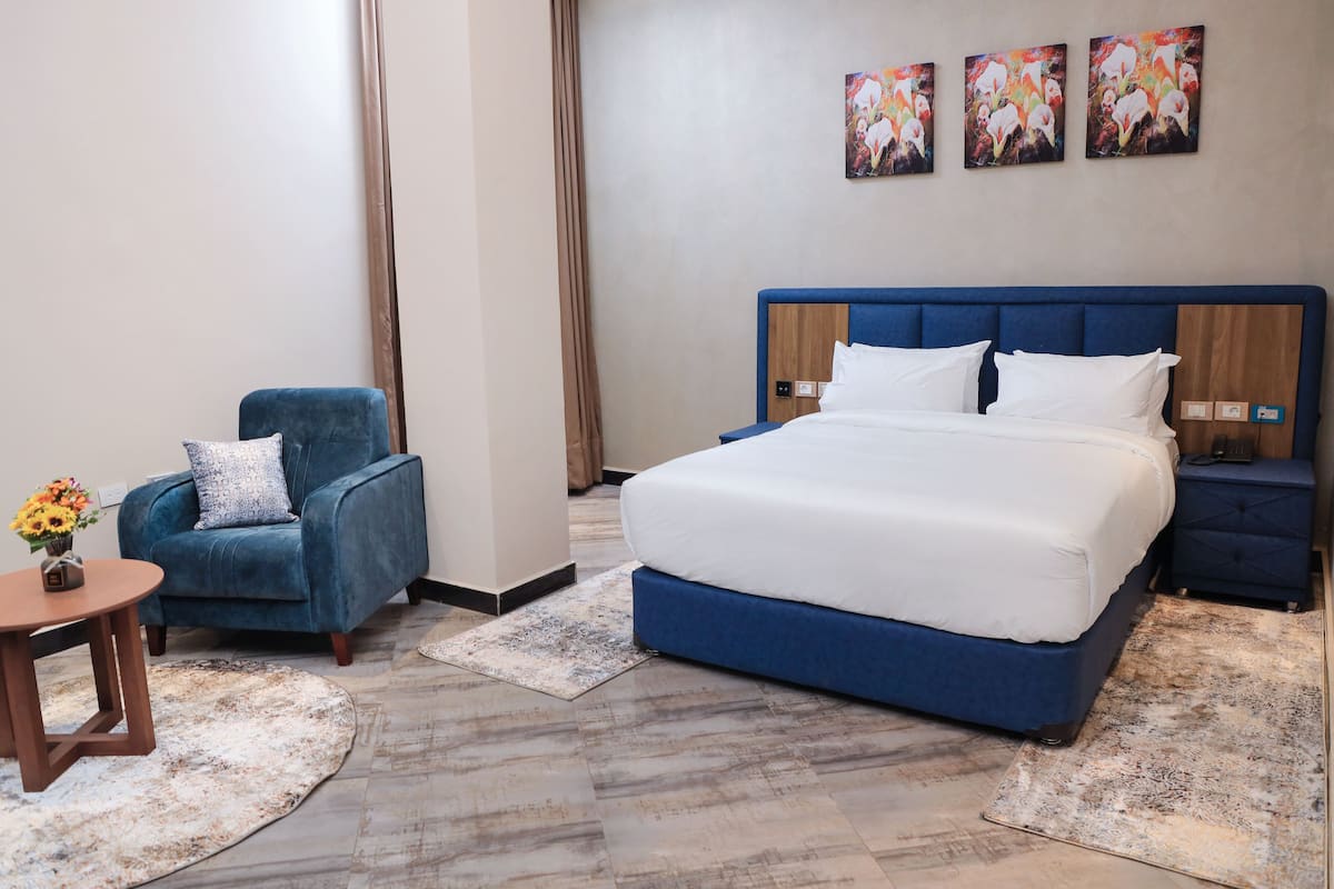 Une chambre d’hôtel avec un grand lit, un fauteuil bleu, une petite table sur laquelle est posé un vase de fleurs, et deux tableaux accrochés au mur.