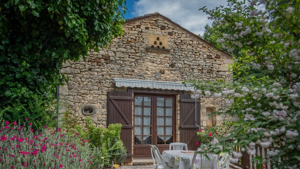 Un edificio in pietra con una porta e delle finestre in legno, immerso in un giardino fiorito e con un vialetto in ghiaia.