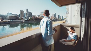 Un hombre de pie en un balcón con vista a un río y edificios al fondo.