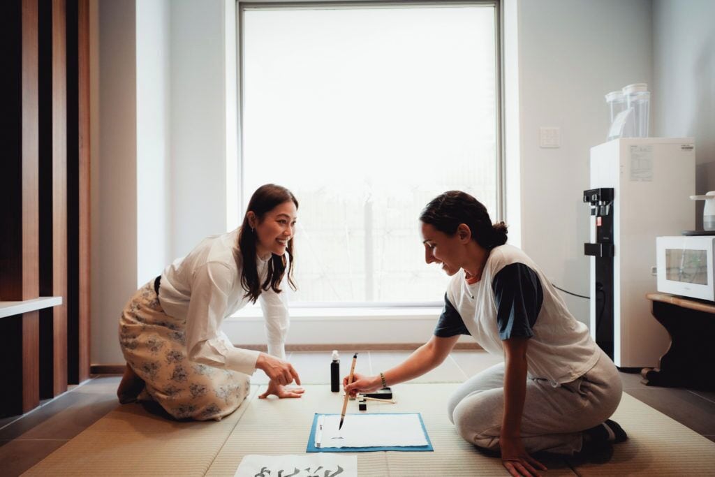 Twee personen zitten op tatami-matten en maken een tekening met papier en kwasten op de vloer.