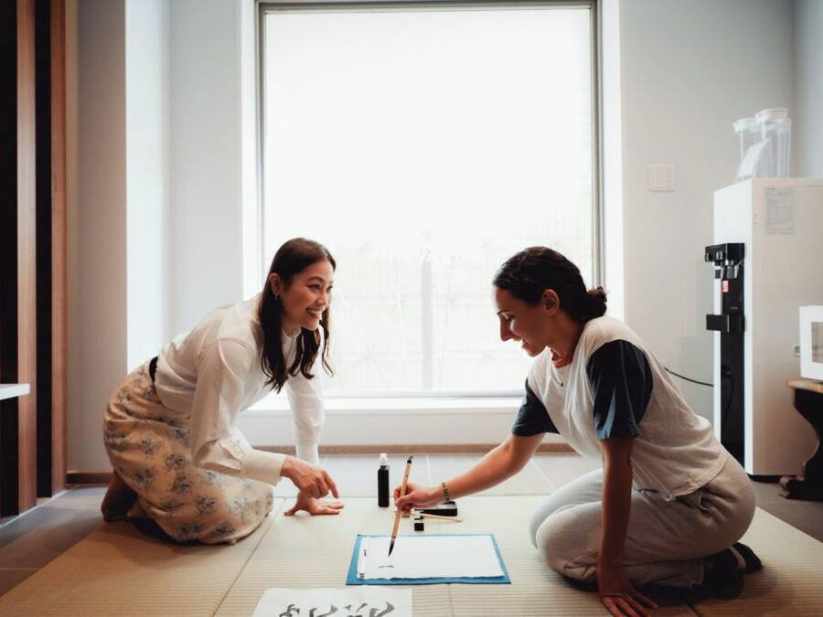 Twee personen zitten op tatami-matten en maken een tekening met papier en kwasten op de vloer.