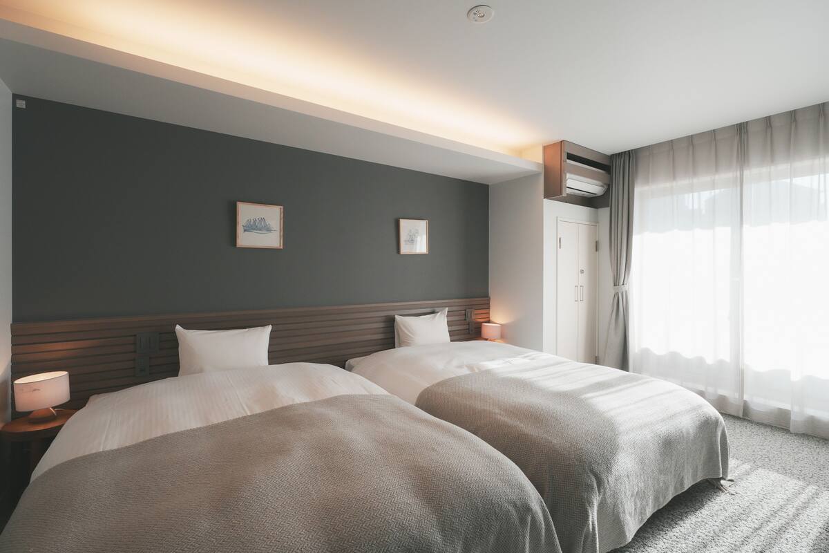 Een hotelkamer met twee bedden, een houten hoofdbord, nachtlampjes en een groot raam met dunne gordijnen.