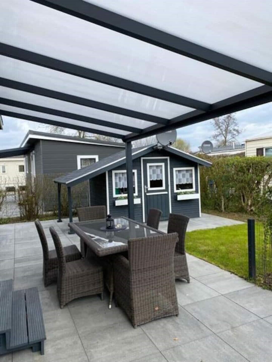 Ein Außenbereich mit einer Terrasse, einem Tisch mit Glasplatte, Korbstühlen, einer Pergola und Blick auf angrenzende Häuser.