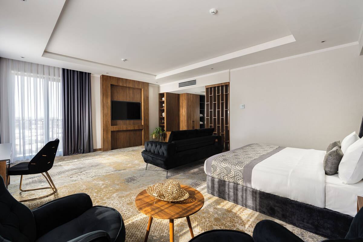A modern hotel room with a large bed, a black sofa, a round wooden table, and a TV mounted on the wall.
