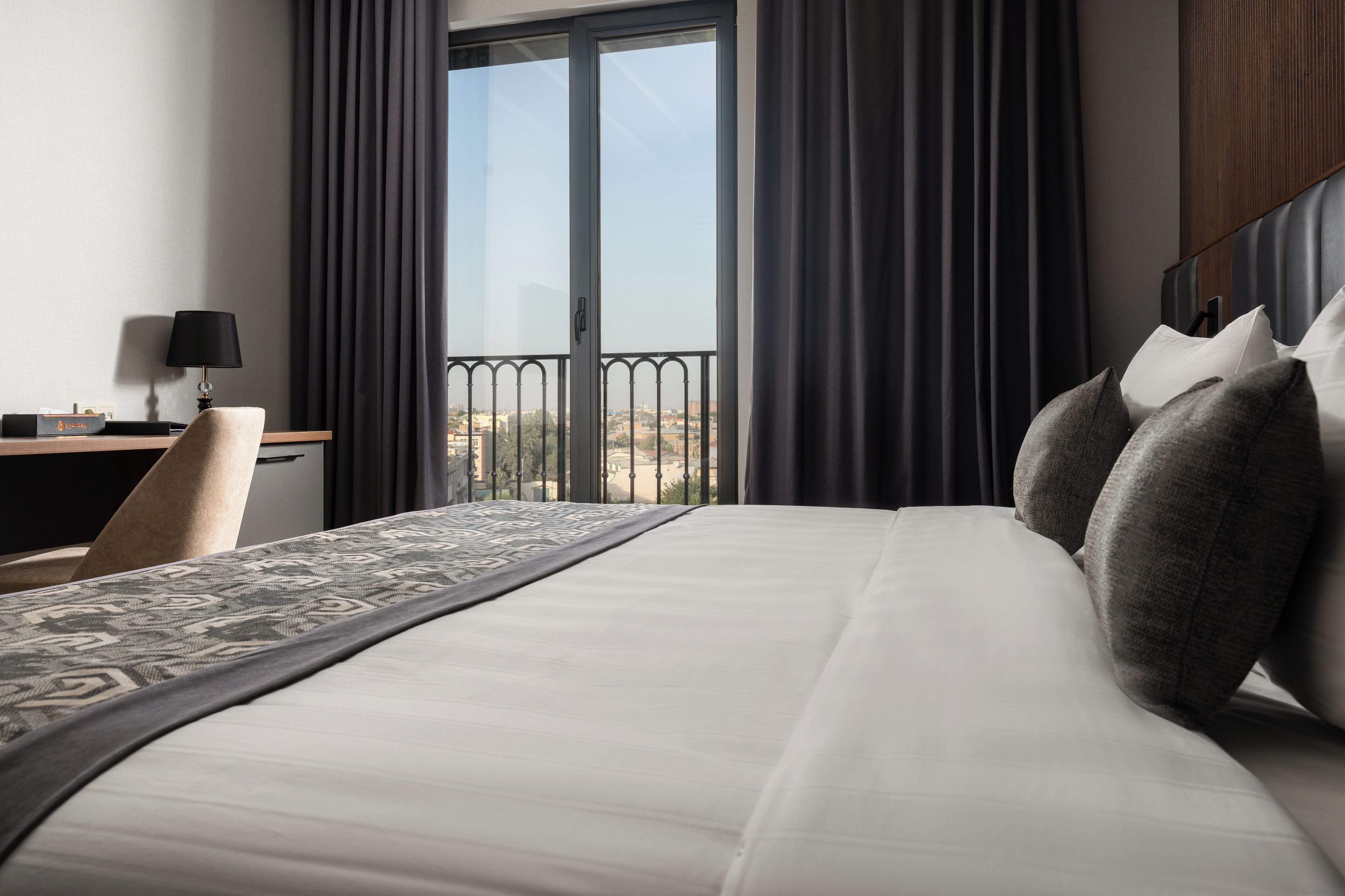 A hotel room with a large bed, a desk with a lamp, and a view of the city through a balcony.