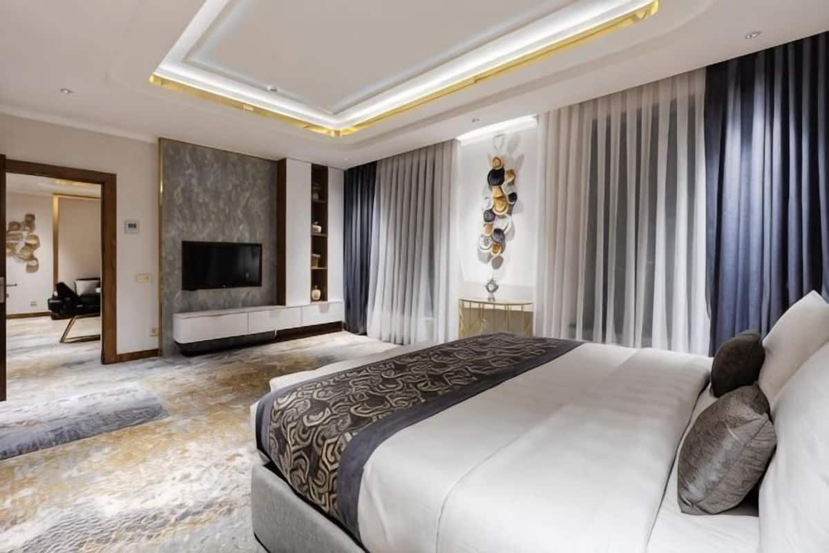 A modern hotel room with a large bed, a flat-screen TV, a seating area with a sofa and armchairs, and a decorative wall piece.