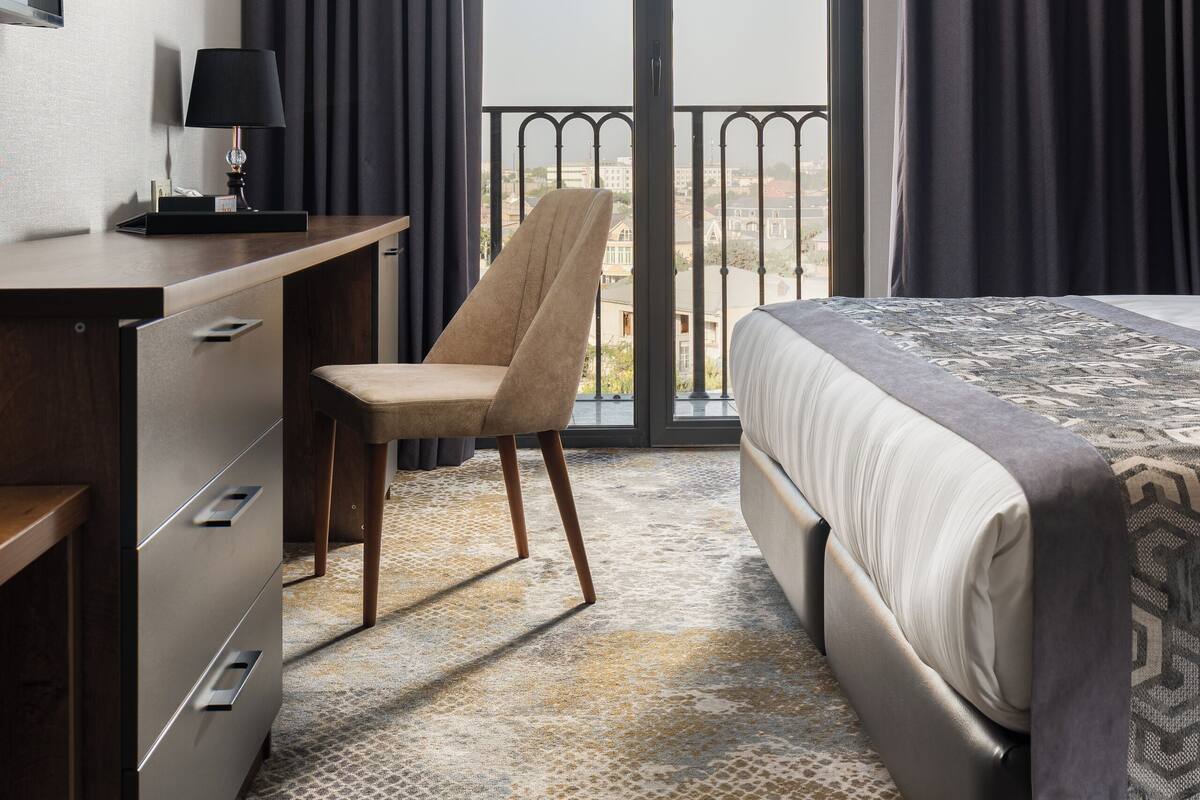 A modern hotel room with a large bed, a desk with a lamp, a chair, and a view of the city through the balcony.