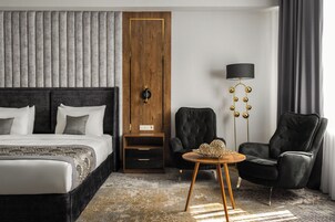 A modern hotel room with a large bed, a wooden wardrobe, a black leather armchair, a small round table, and a floor lamp.