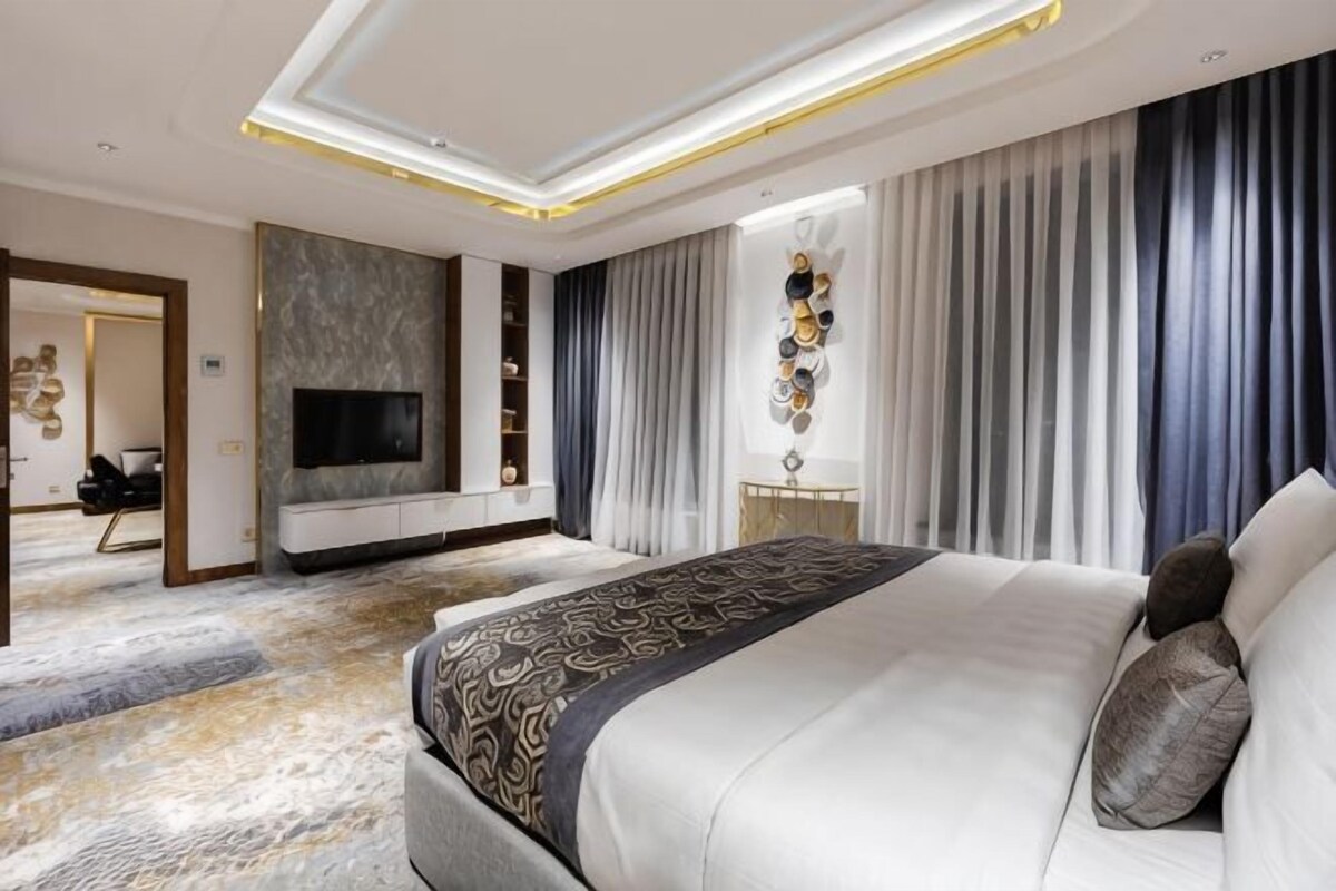 Une chambre d’hôtel moderne dotée d’un grand lit, d’un téléviseur à écran plat, d’un coin salon avec un canapé et des fauteuils, et d’un élément décoratif mural.