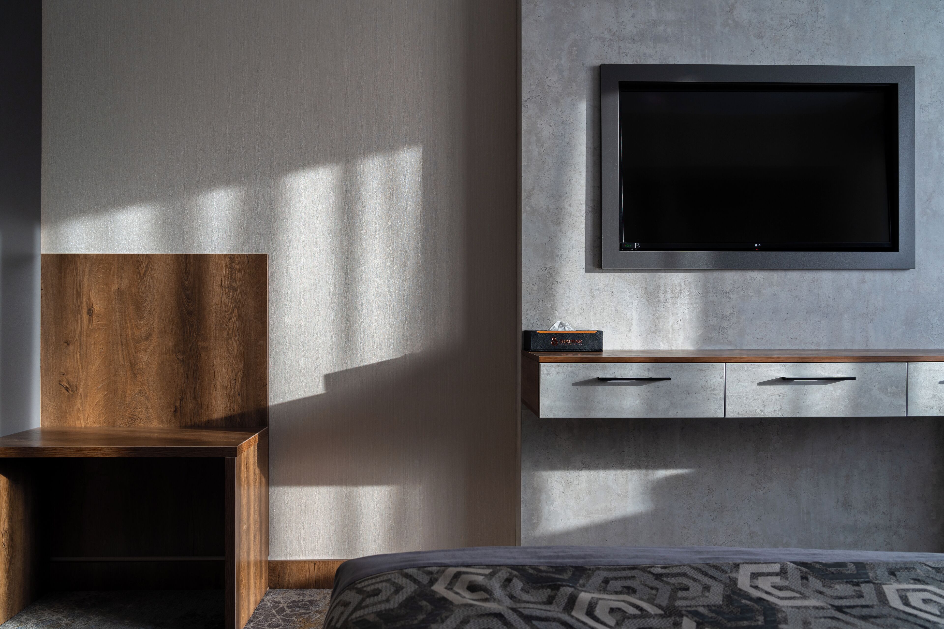 Ein modernes Hotelzimmer mit einem großen Flachbildfernseher an der Wand, einem hölzernen Nachttisch, einer bettenbezogenen Auflage mit Muster und einer eingebauten Stauraum-Einheit mit Schubladen.