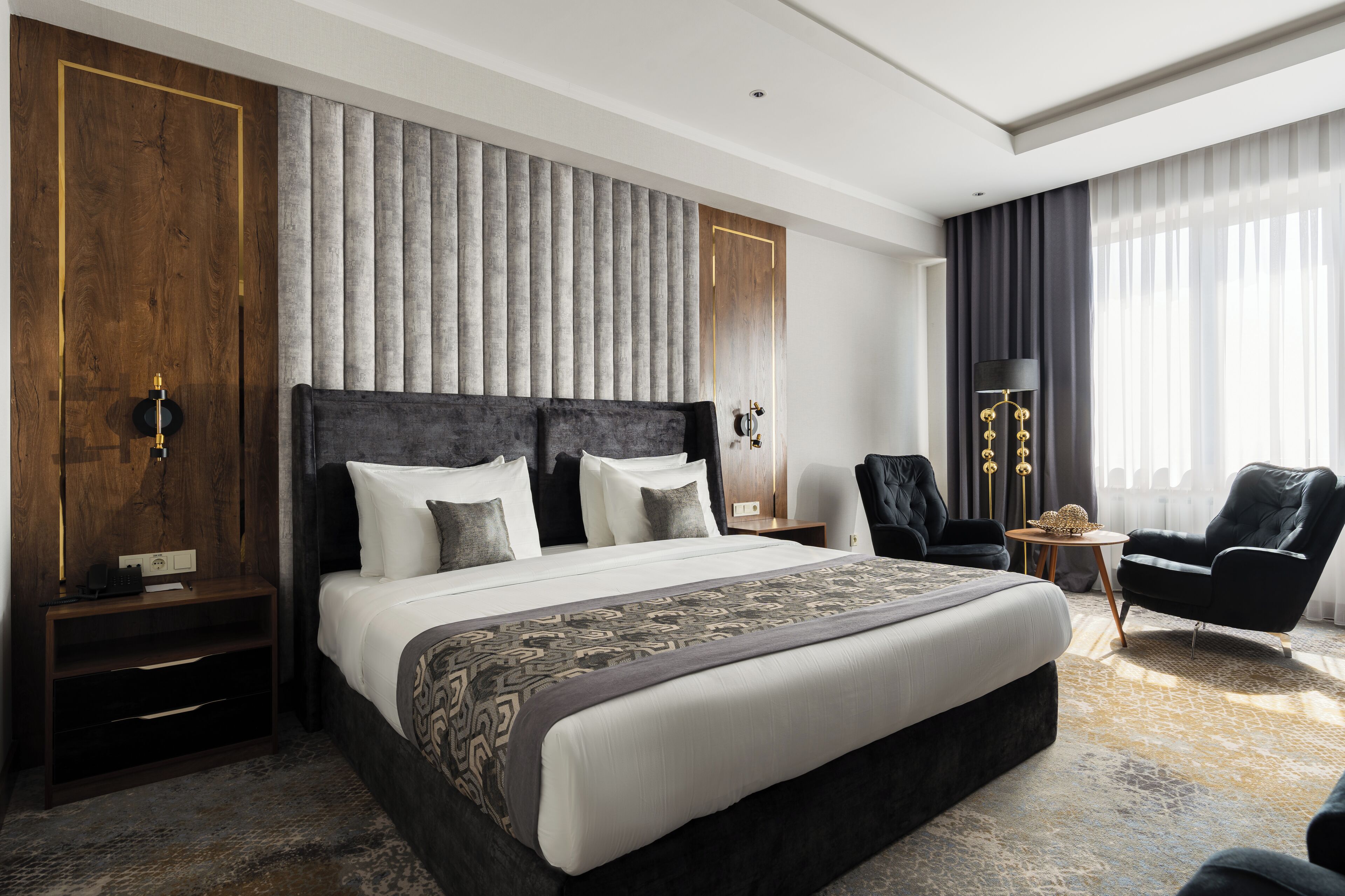 A modern hotel room with a large bed, a nightstand, a chair, a small table, and a lamp.