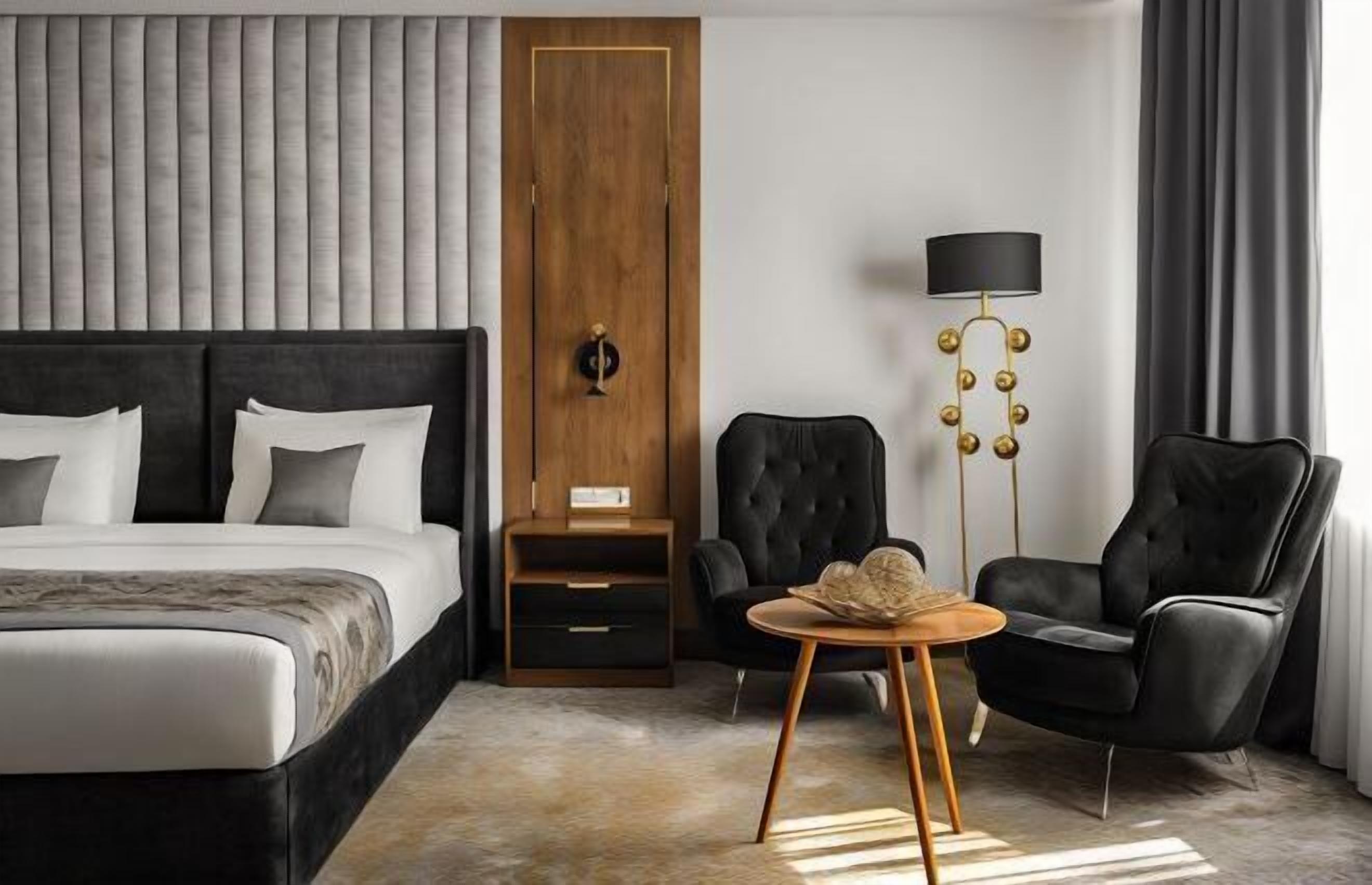 A modern hotel room with a large bed, a wooden wardrobe, a black armchair, a round wooden table, and a floor lamp.